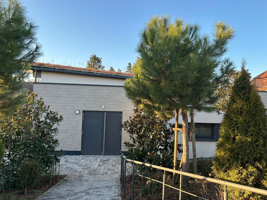 Außenansicht eines modernen, flachen Gebäudes mit hellbeiger Fassade in Holzoptik und anthrazitfarbenen Doppeltüren. Im Vordergrund führt ein gepflasterter Weg zum Eingang, flankiert von grünen Nadelbäumen und Sträuchern unter einem klaren blauen Himmel.